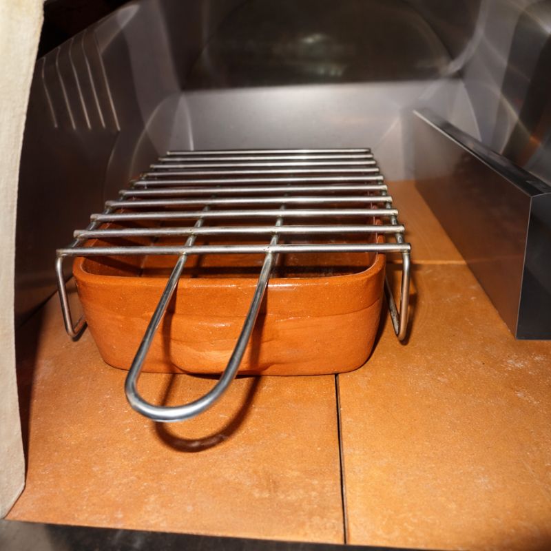 BBQ rack positioned over a terracotta baking dish inside a wood-fired pizza oven