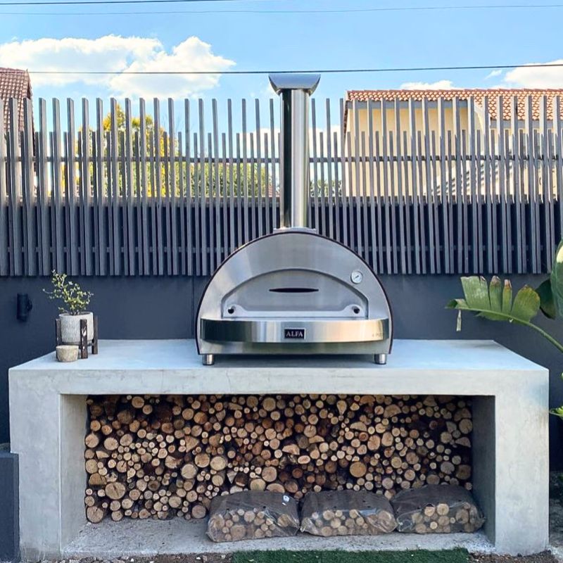 Alfa 4 Pizze Wood Pizza Oven on a concrete platform with stacked logs underneath, against a blue sky | Premium Pizza Oven Sale