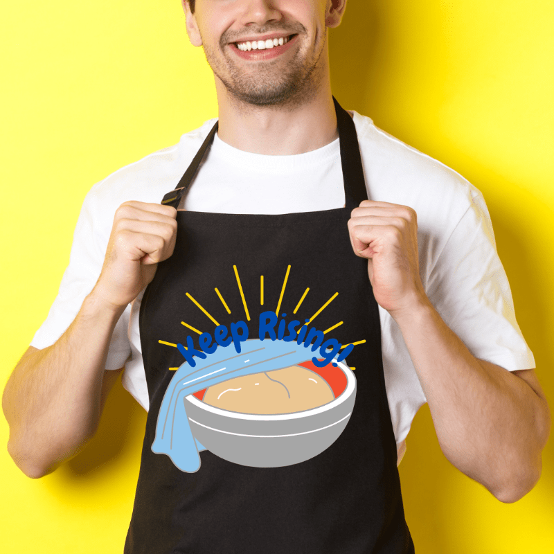 upclose photo of black apron featuring the design "proofing bowl with phrase keep rising" on it 