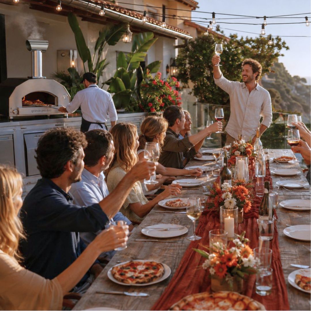 A group of people gathered around a long dining table outdoors at sunset in the foreground, and the ItaliaForno Grande Pro Dup Hybrid Pizza Oven sooking in the background