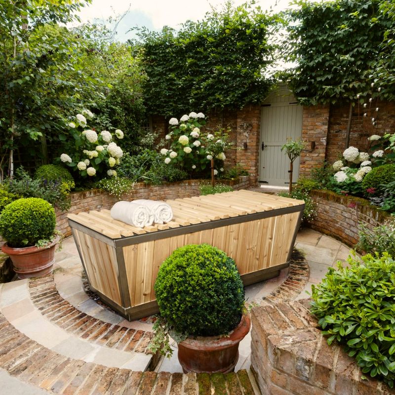 LeisureCraft Canadian Timber Polar Plunge Tub in a landscaped backyard, surrounded by brickwork, hydrangeas, and trimmed bushes.